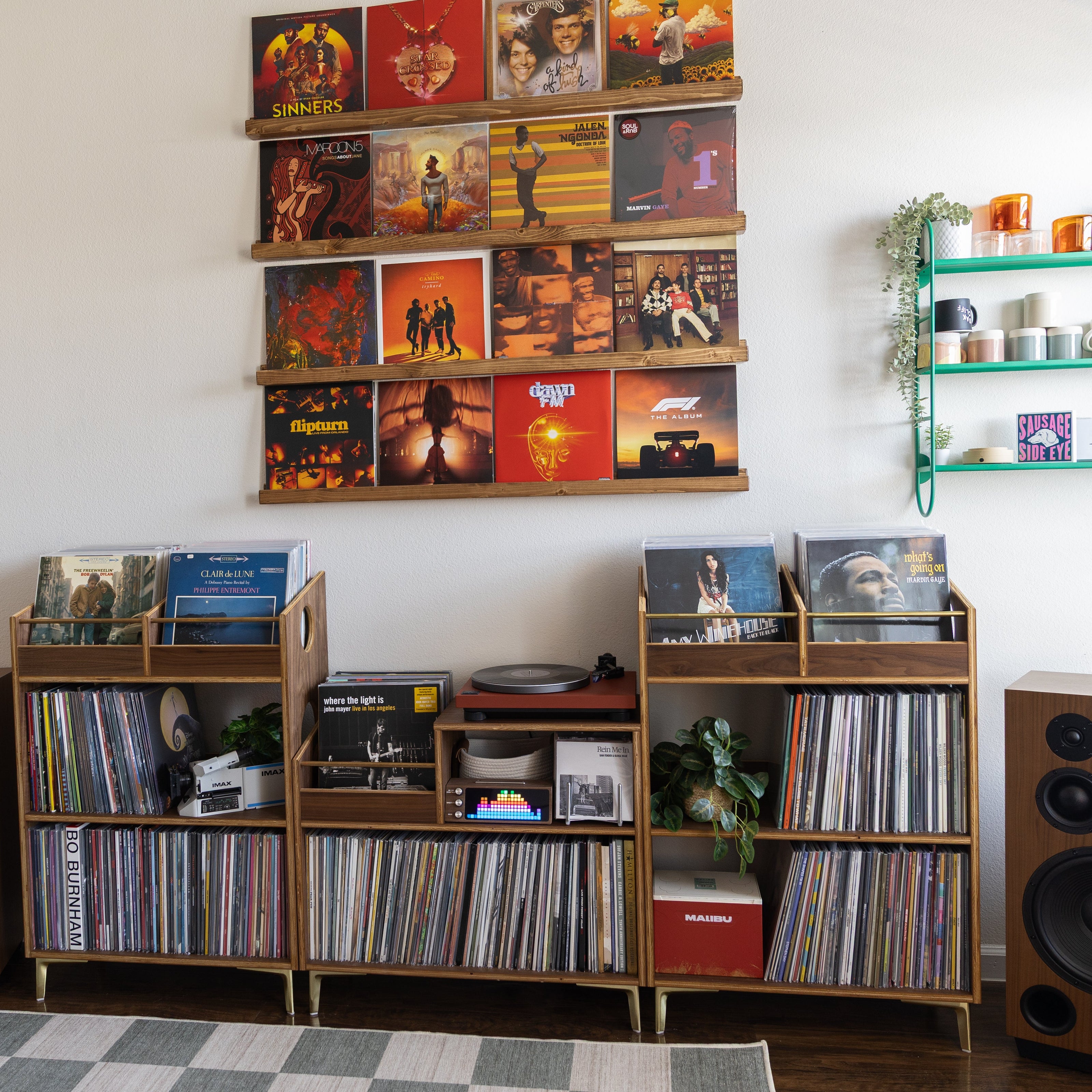 A styled home audio room featuring three mid-century modern wooden record shelves filled with vinyl records, flanked by two large wooden speakers. Above the shelves, a wooden floating shelf unit holds a grid of colorful album covers, and a green wire wall shelf with plants is mounted on the white wall.