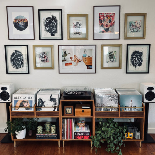 Music collection with vinyl records and speakers on a wooden shelf against a wall with framed artwork.