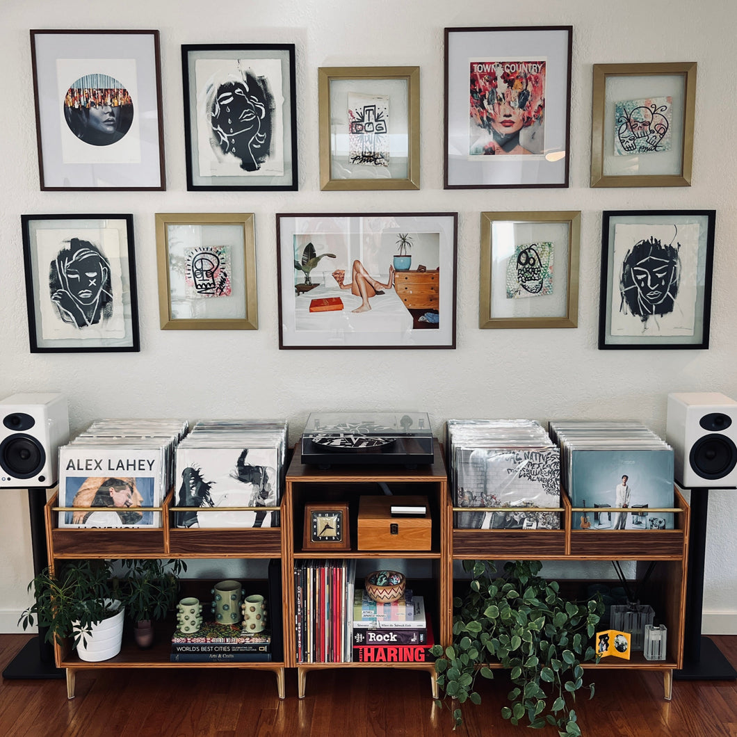 Music collection with vinyl records and speakers on a wooden shelf against a wall with framed artwork.