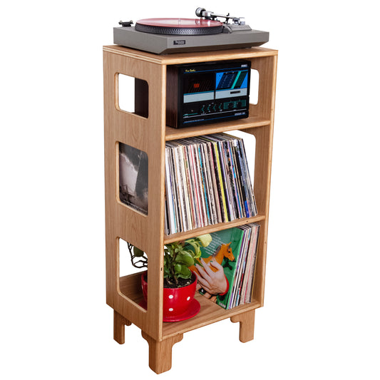 A light wood record player stand with three open shelves, holding a turntable on top, a stereo receiver, vinyl records, a small plant, and a red vase, isolated on a white background.