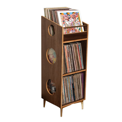 A mid-century modern wooden record player stand with three open shelves holding vinyl records and a record sleeve, featuring circular cutouts on the side and tapered legs, isolated on a white background.