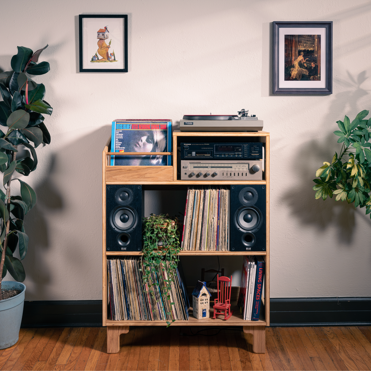 A mid-century modern wooden record shelf cabinet styled in a room corner with potted plants, framed wall art, and a hardwood floor. The cabinet features open shelves for records, a side shelf for a turntable and cassette deck, and built-in speaker compartments.