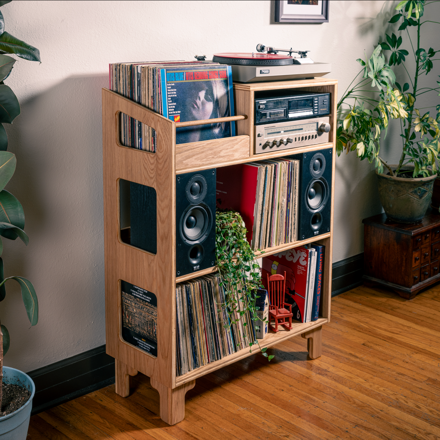 A light natural wood record shelf and speaker cabinet stands on a hardwood floor in a styled room, filled with vinyl records, speakers, a turntable, and small decor items. Large potted plants and a dark wood side table are visible in the background.