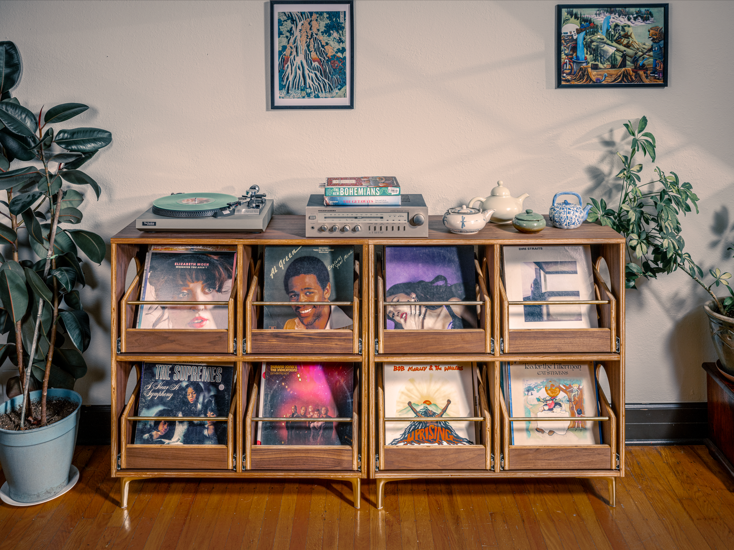 Wooden record player console with vinyl records in a room with plants and framed pictures.