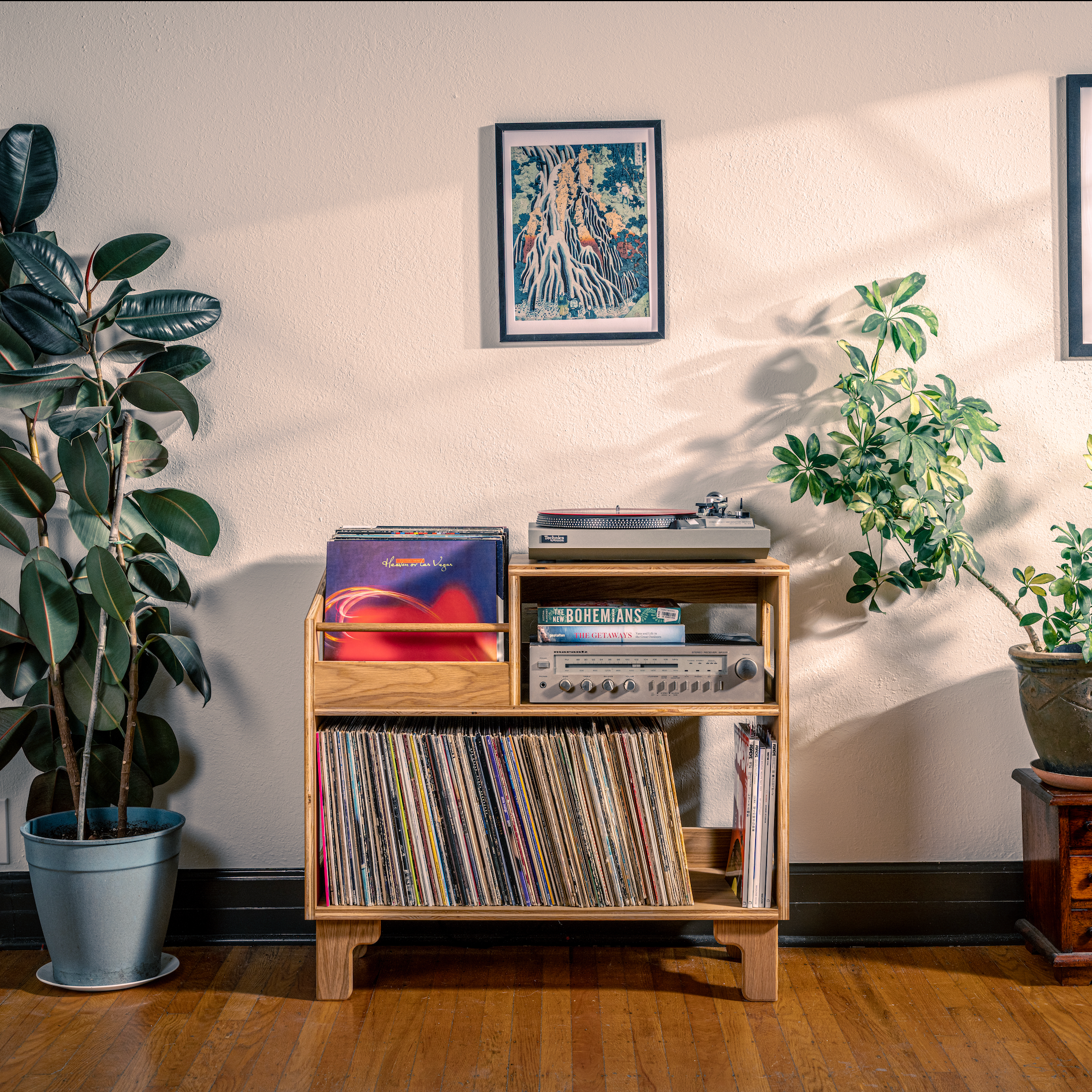 Wooden record player with vinyl records, surrounded by potted plants and framed posters on a wall.