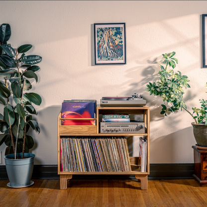 Wooden record player with vinyl records, surrounded by potted plants and framed posters on a wall.