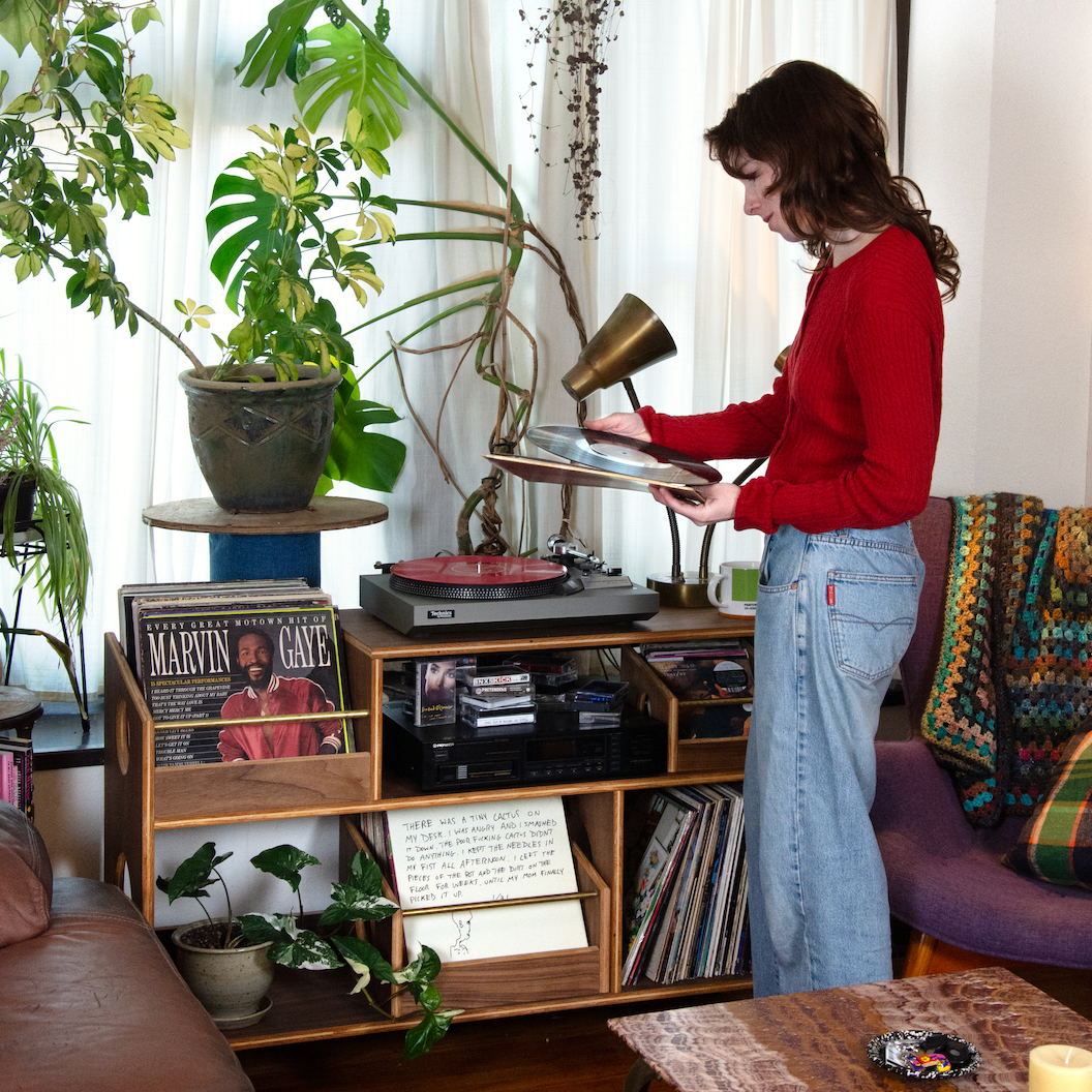Woman playing a vinyl record in a cozy living room with plants and a record player.