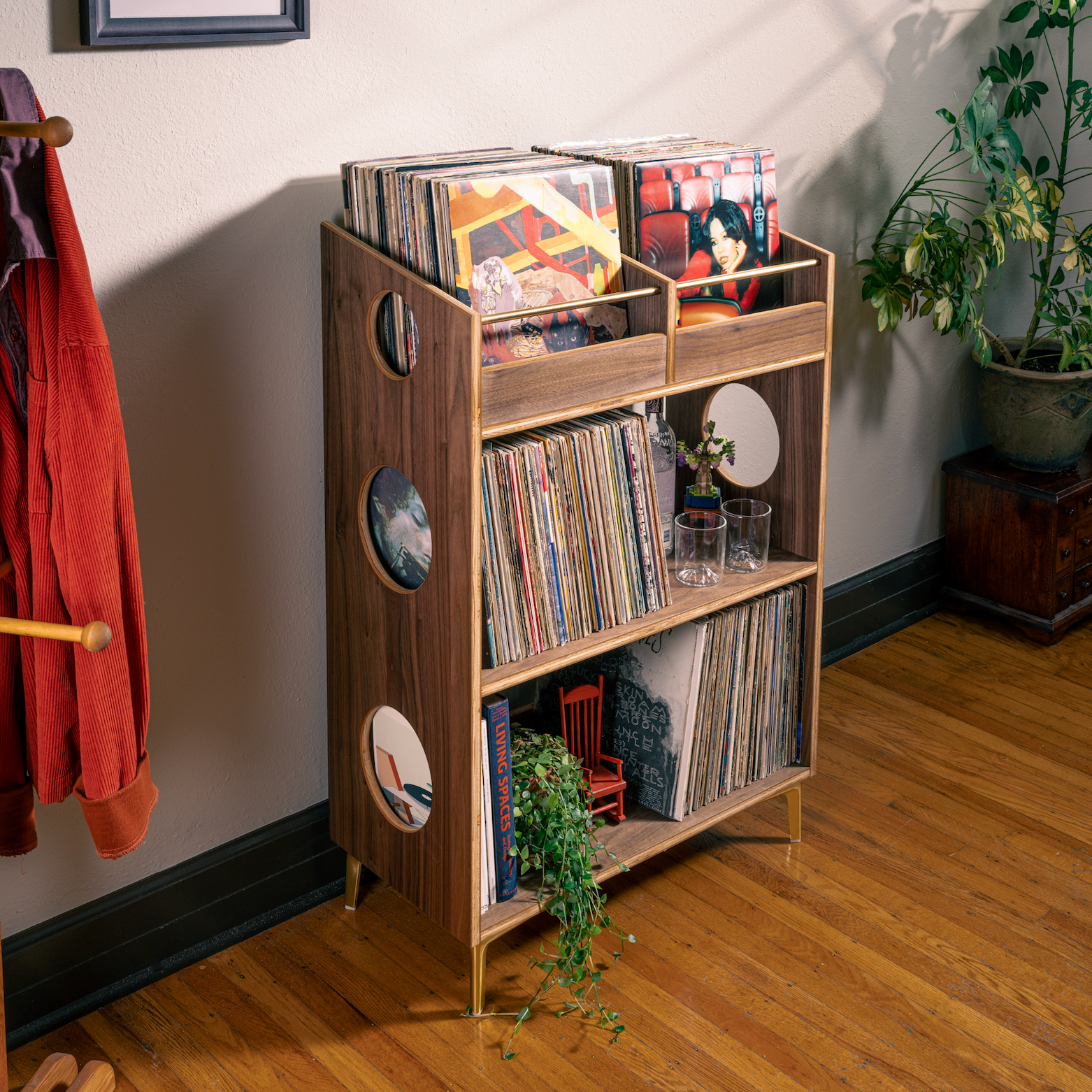 A mid-century modern wooden record shelf with gold legs, styled in a cozy room corner with plants, a coat rack, and vinyl records.