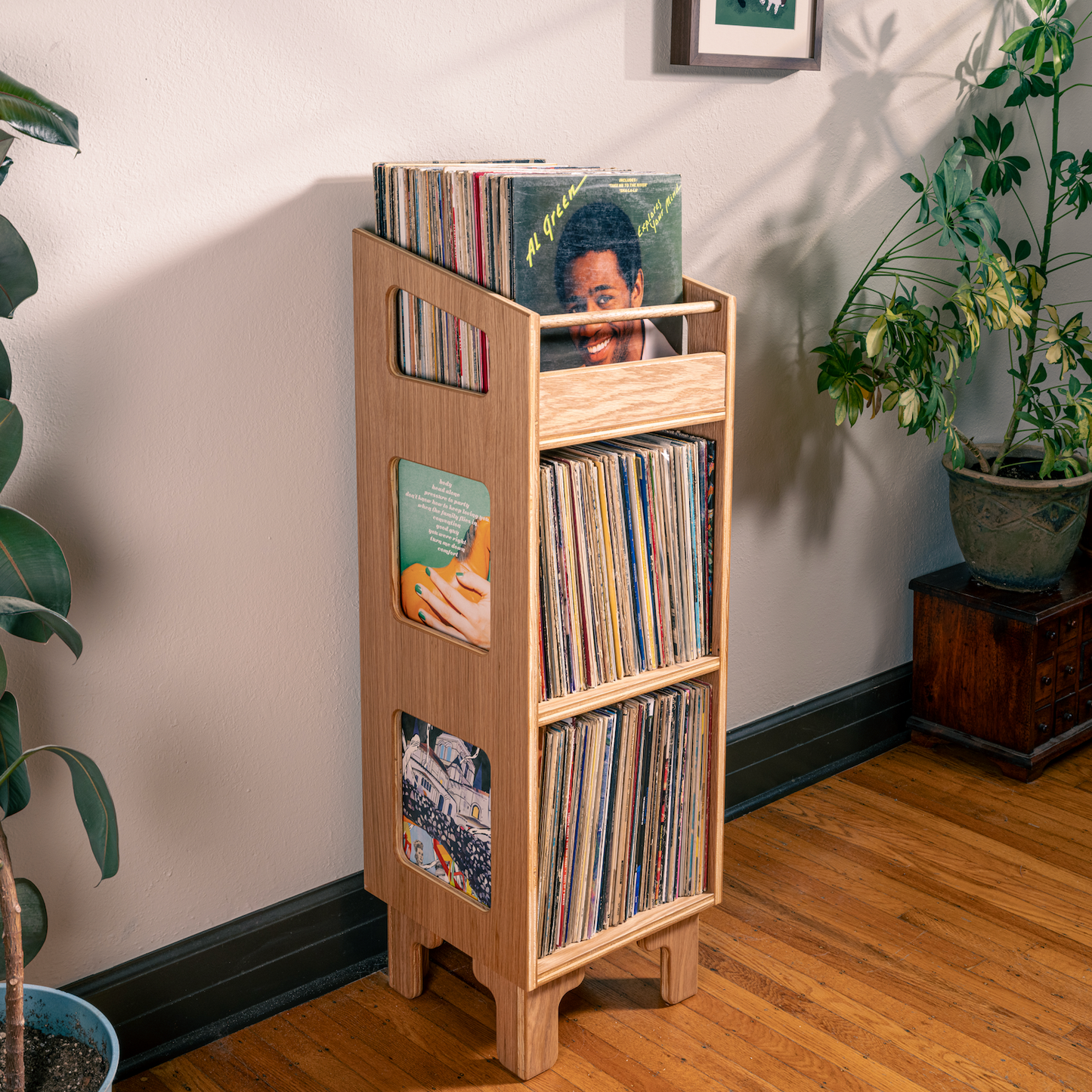 A light natural wood record rack with three tiers stands on a hardwood floor in a room corner, holding vinyl records and album covers. Large potted plants and a potted tree on a wooden stand are visible against a beige wall.