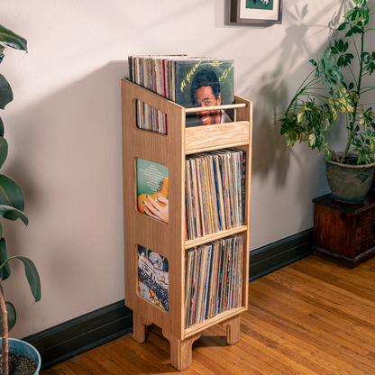 A light natural wood record rack with three tiers stands on a hardwood floor in a room corner, holding vinyl records and album covers. Large potted plants and a potted tree on a wooden stand are visible against a beige wall.
