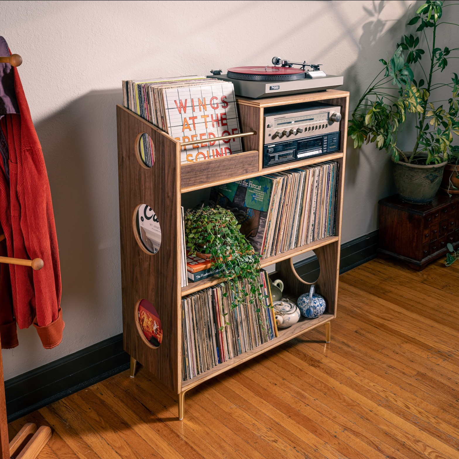 A mid-century modern wooden record shelf with gold legs stands in a room corner on a hardwood floor, filled with vinyl records, a turntable, and decorative plants.