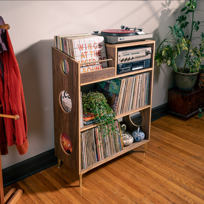 A mid-century modern wooden record shelf with gold legs stands in a room corner on a hardwood floor, filled with vinyl records, a turntable, and decorative plants.
