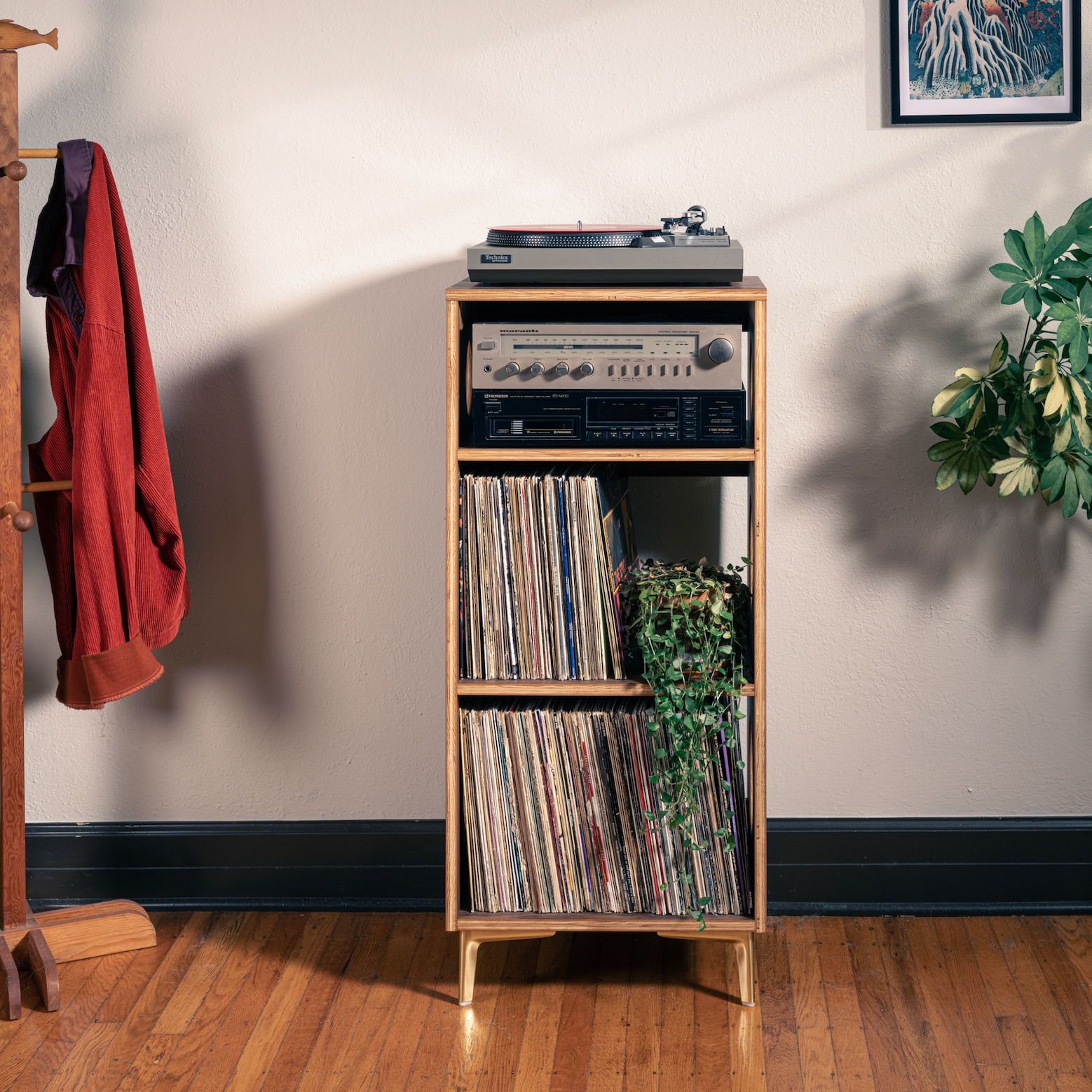 A wooden record cabinet with open shelves stands on a hardwood floor against a beige wall, holding vinyl records and a turntable. A coat rack with a red jacket is on the left, and potted plants are on both sides.