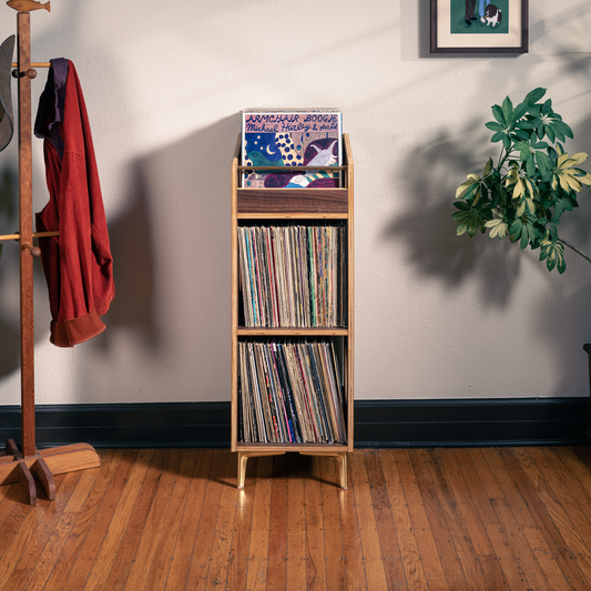 A tall wooden record rack with three shelves stands against a light wall in a styled room. The top shelf holds a vinyl record, and the middle and lower shelves are filled with records.