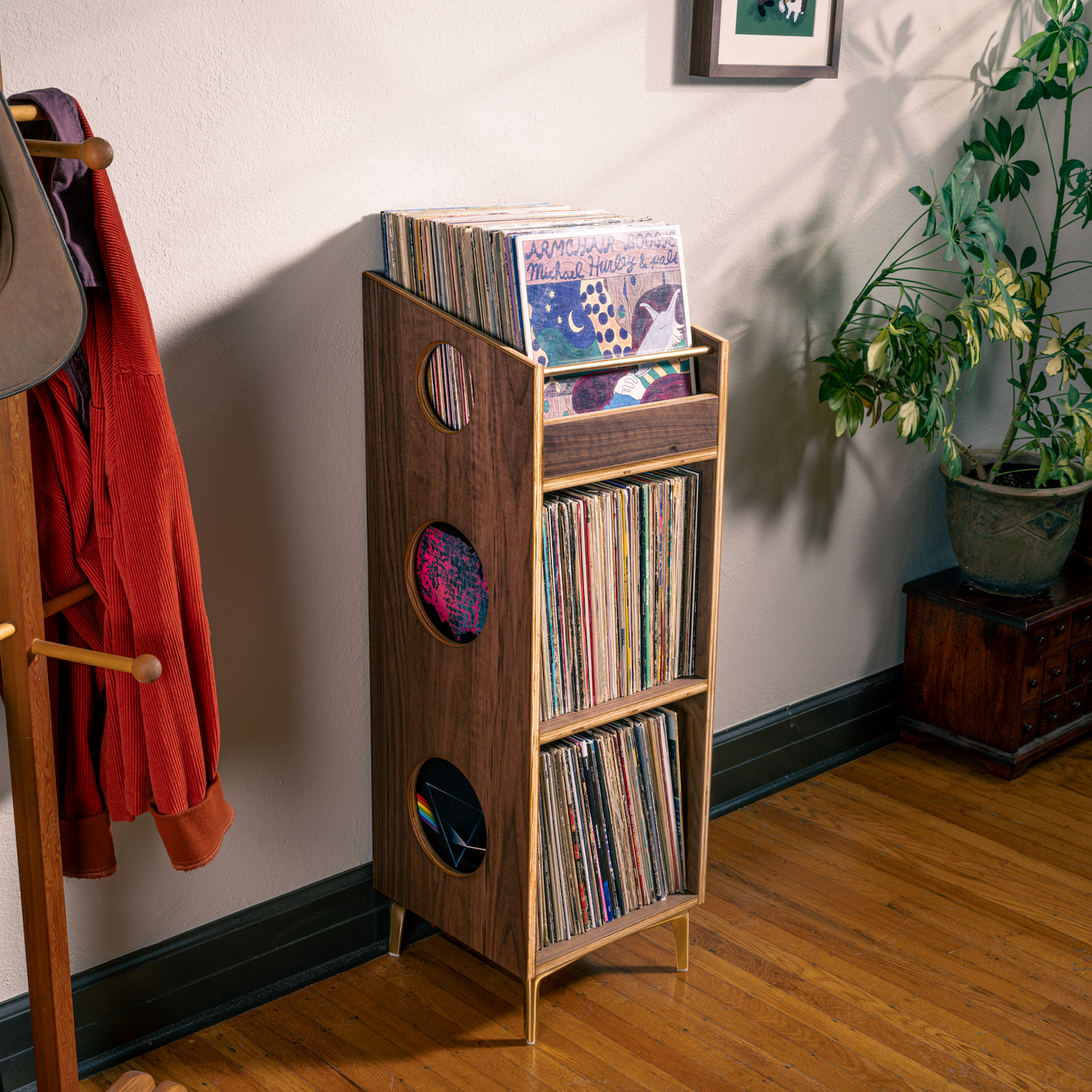 A tall wooden record rack with three shelves and circular cutouts, holding vinyl records, placed in a room corner with potted plants, a coat rack, and a hat nearby.