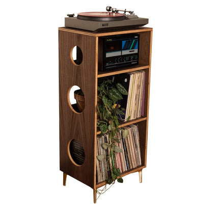 A mid-century modern wooden record player cabinet with gold legs, featuring a turntable on top, a small display screen, and shelves holding vinyl records and greenery, isolated on a white background.