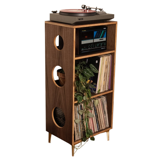 A mid-century modern wooden record player cabinet with gold legs, featuring a turntable on top, a small display screen, and shelves holding vinyl records and greenery, isolated on a white background.