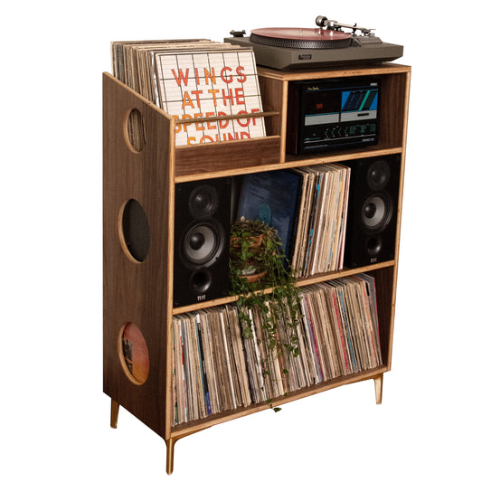 A mid-century modern wooden record shelf and speaker cabinet with a turntable on top, filled with vinyl records, flanked by two speakers, on a plain white background.